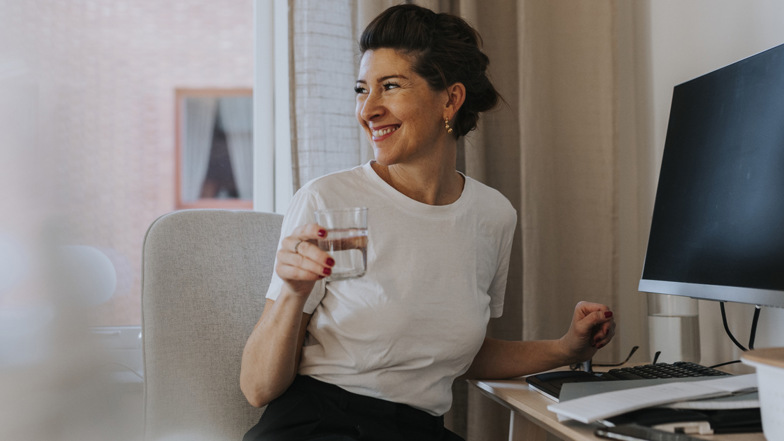 En leende kvinna vid ett skrivbord med dator och blanketter håller i ett vattenglas och vänder sig mot rummet. 