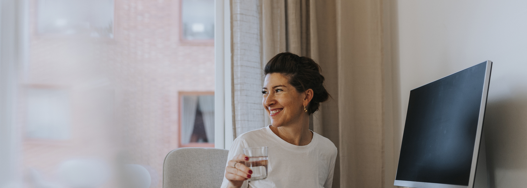 Mörkhårig leende kvinna iklädd vit t-shirt med uppsatt hår sitter bortvänd från en dator i ett hem med ett glas i handen. 