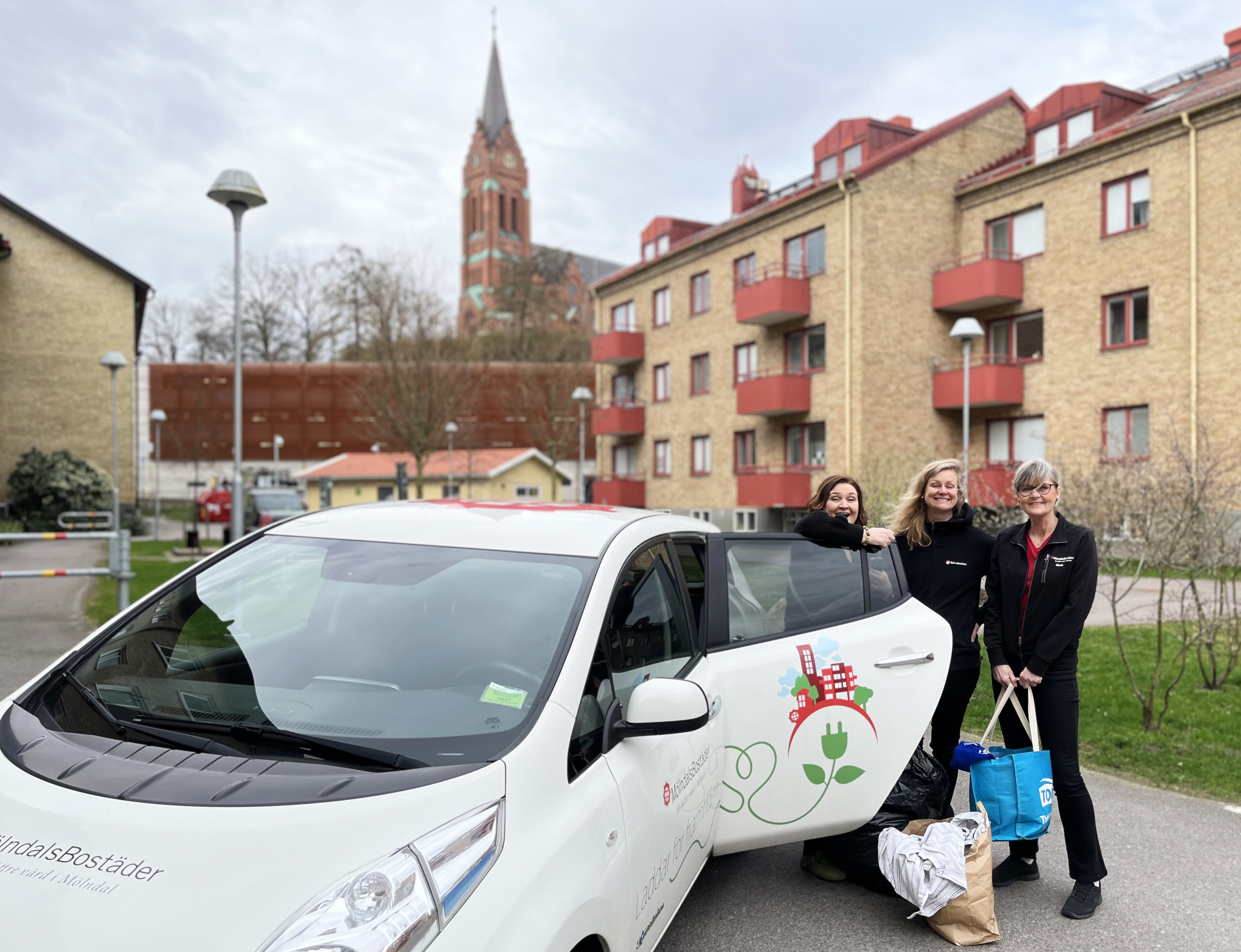 Tre kvinnor står leende och poserar bredvid en bil med öppen bakdörr i ett hyreshusområde med Fässbergs kyrka i bakgrunden. 