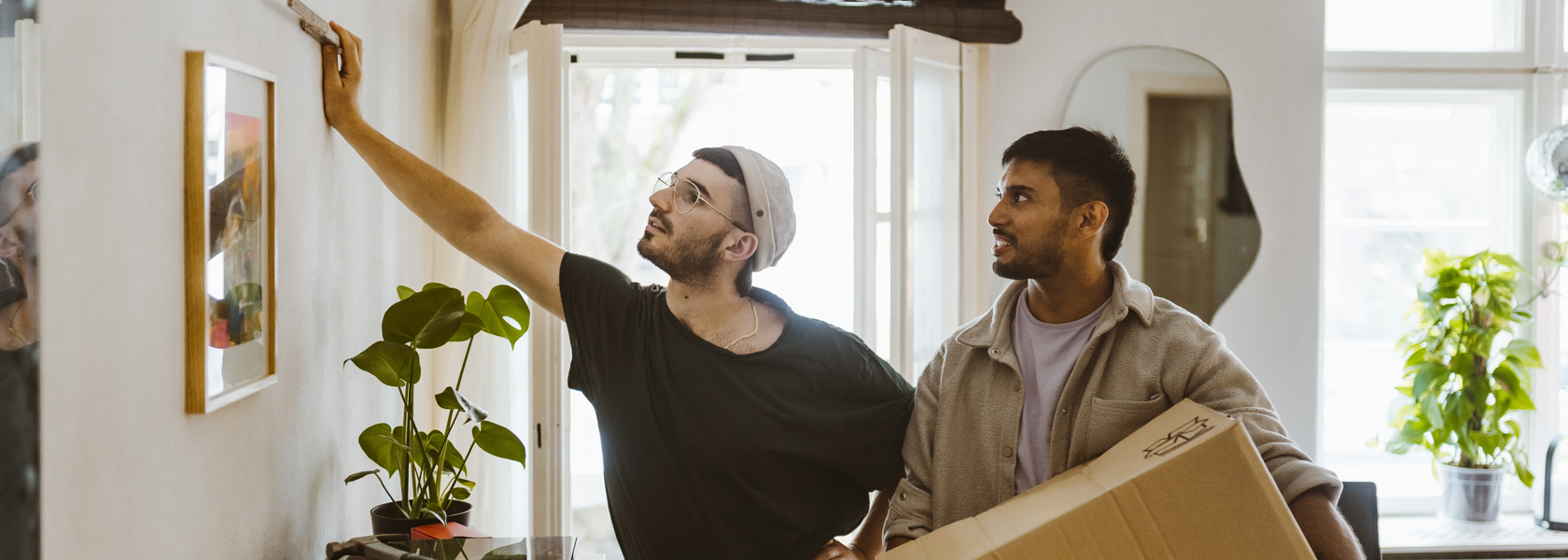 Två män i en lägenhet där den ena mäter på en vägg och den andra håller en flyttkartong och tittar på.